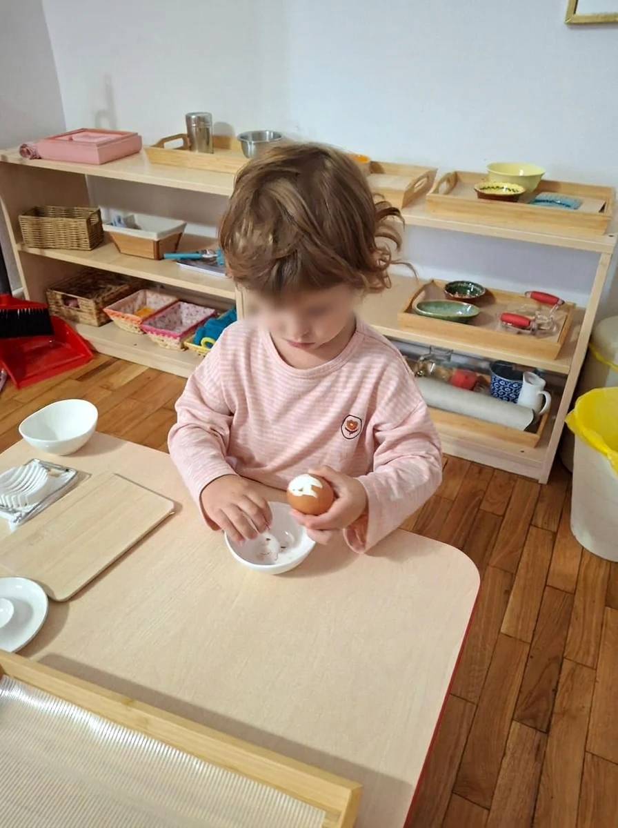 Copil spărgând un ou la masă, cu mobilierul și materialele Montessori la înălțimea lui, în Grupa 1,8-3 ani din Arcadia Montessori