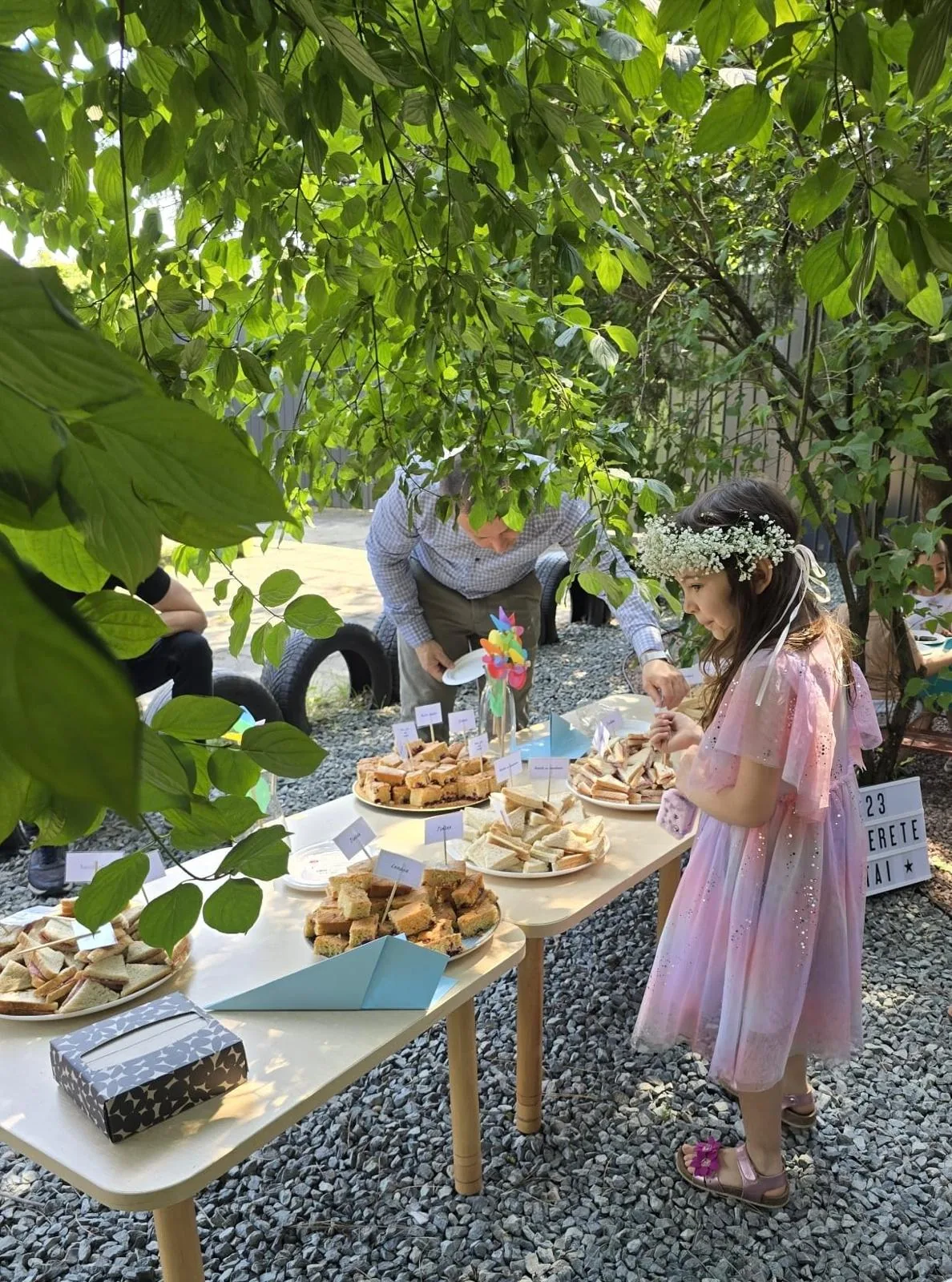 Copil cu coroniță de flori lângă mesele cu gustări, la evenimentul de sfârșit de an școlar 2025–2026, Arcadia Montessori